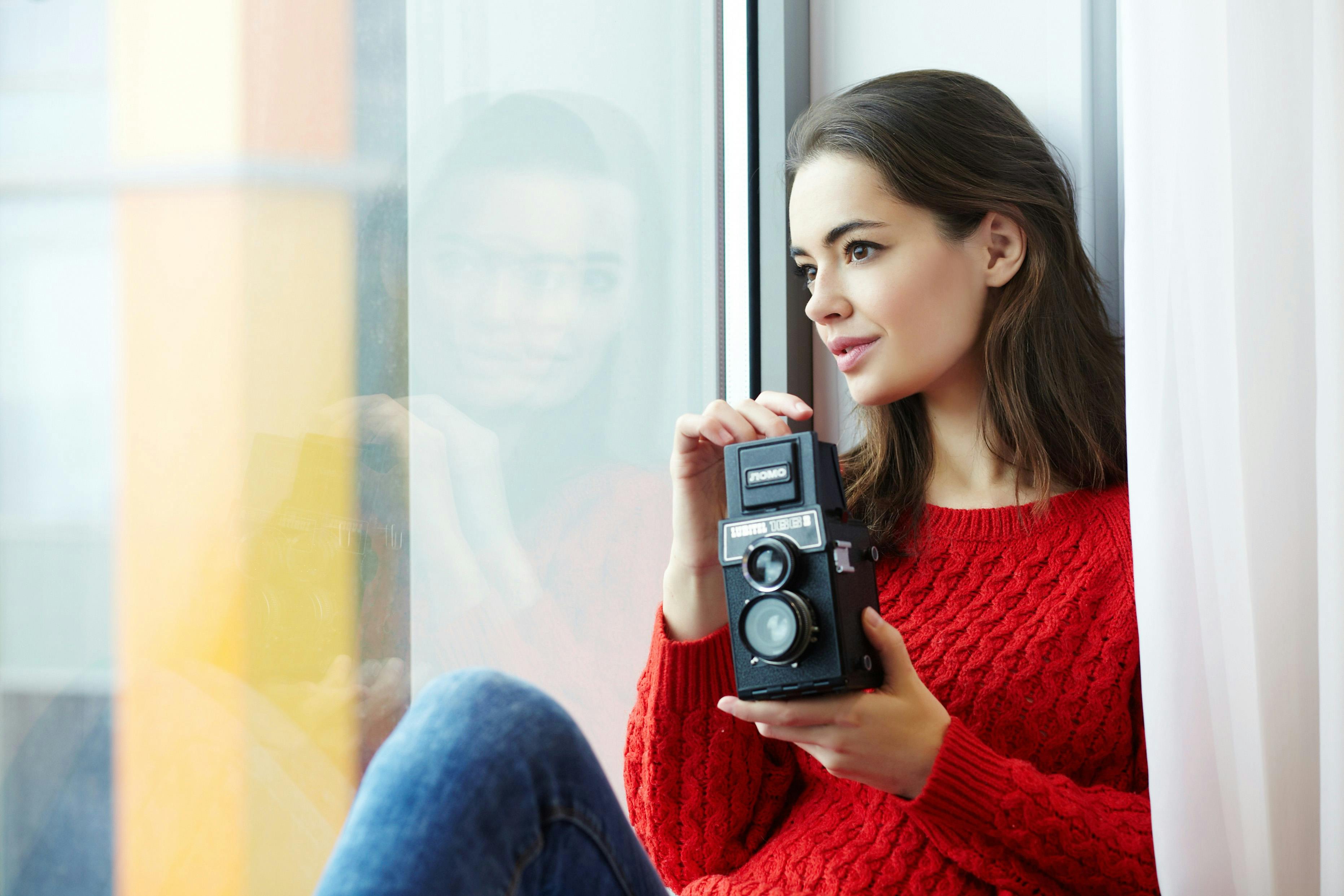 Young woman in a red sweater holding a vintage camera by a window, looking thoughtful.